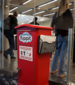 Photo of red Flippit Dropbox outside of the Pre-Board Screening Checkpoint at YQR Regina International Airport. In the background there are passengers waiting in the queue to be screened.